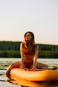 Young woman in red bikini sits on a paddle board on a calm lake during the warm golden sunset hours