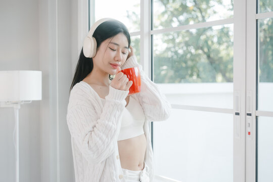 Young woman in white loungewear sits by a bright window, wearing headphones and drinking from a red mug in a calm modern home setting
