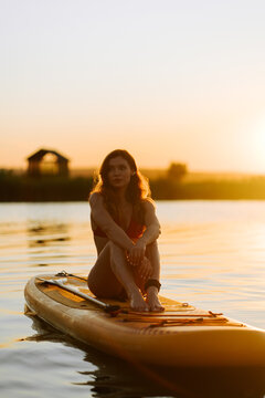 Young woman sitting on a paddle board in the middle of a calm lake during a golden sunset period