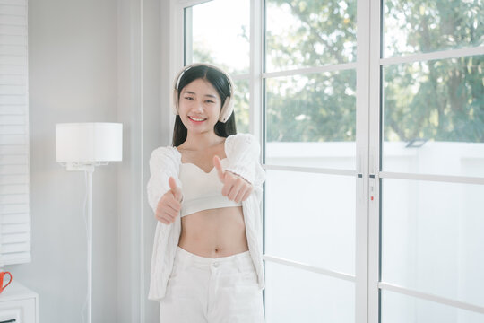 Young woman in white outfit wearing headphones and standing by a bright window at home, smiling with folded arms in a calm modern interior.