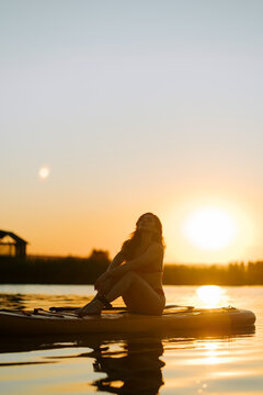 Woman sitting on a stand up paddle board relaxing on a quiet lake during a beautiful golden sunset
