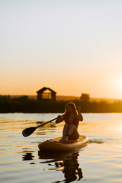 Young woman sitting on a paddle board while paddling across a calm river during a golden sunset