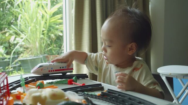 A young child engages with a colorful toy train set on a table beside a window, surrounded by greenery. The scene captures playful curiosity and the joy of imaginative play in a bright and inviting sp