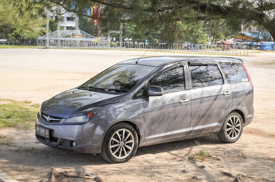 BATAM, INDONESIA - AUGUST 2, 2026: A grey Proton Exora MPV car parked under pine trees on a sandy beach area in Batam, Riau Islands, Indonesia.