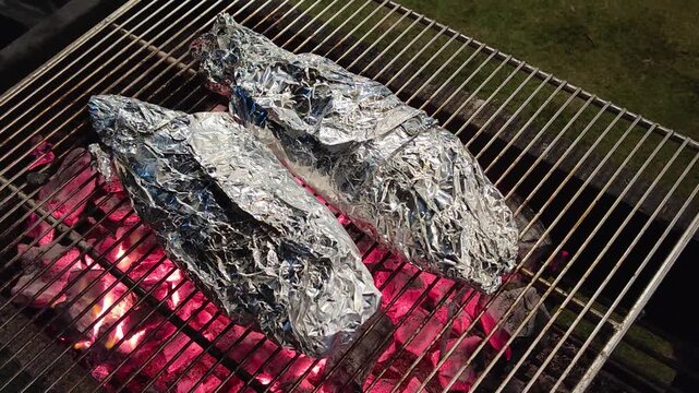 charcoal grilled fish wrapped in aluminum foil cooking over open fire during outdoor bbq, smoky and juicy summer meal