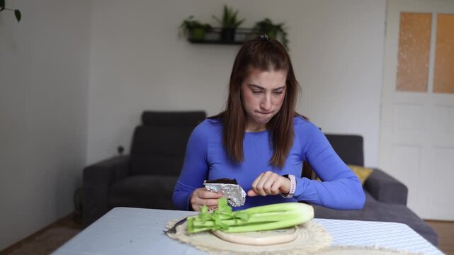 Woman struggles to choose between healthy celery and tempting chocolate showing the daily battle between nutrition goals cravings and self control at home