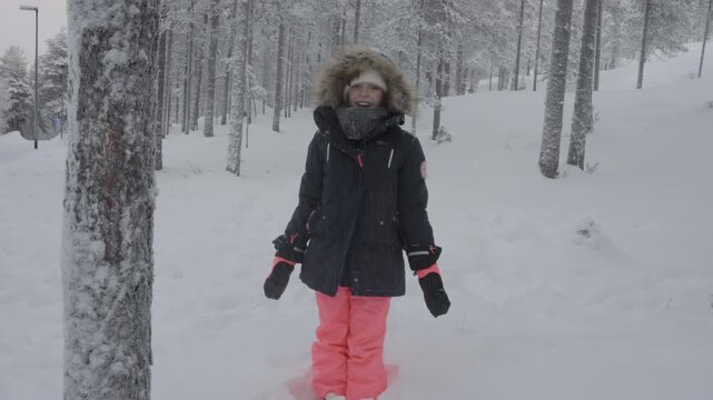 A happy child stands in a snow covered forest while heavy snow falls down upon her in winter. She wears a warm parka with a fur lined hood, colorful ski pants, and gloves outdoors