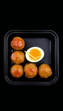 A black container holding five glazed meatballs and a soft boiled egg garnished with thin cheese