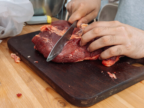 Hands cutting raw beef into smaller pieces on a cutting board before grinding meat in home kitchen.