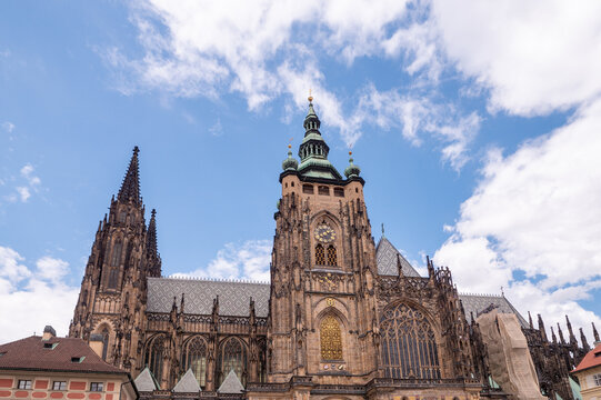 St. Vitus Cathedral at Prague Castle in Prague, Czechia, with its Great South Tower, historic clock and ornate Gothic architecture under a bright blue sky with white clouds.