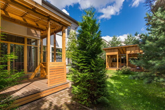 The scene shows a wooden terrace next to a garden with trees and grass. A gazebo is visible in the background. The sky is partly cloudy and bright. It is daytime