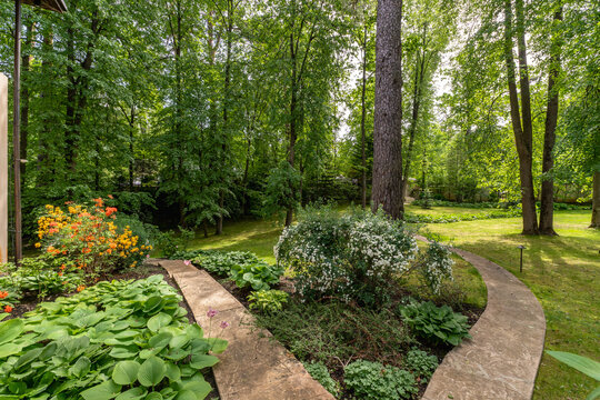 A winding pathway leads through a garden filled with various plants and flowers. Tall trees provide shade in the background. It is daytime, and the sky is clear