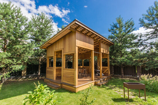 A wooden structure sits in a garden on a sunny day. It has open sides and small steps leading inside. Trees surround the space. A grill is nearby in a green area