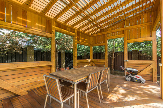 A wooden gazebo stands in a backyard. Inside, there is a table with chairs arranged around it. A lawn mower is parked nearby. Trees and a fence surround the area