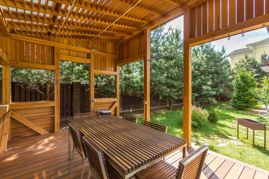 The gazebo has a long wooden table and chairs under a wooden roof. Green trees surround the area, and sunlight enters through the openings. The setting is calm and inviting