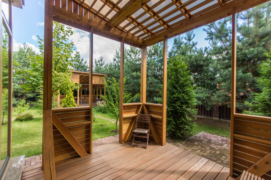 A wooden gazebo stands in a garden. It has a view of green grass and trees. Sunlight filters through the structure. A single chair sits on the wooden floor