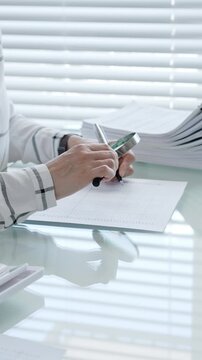Financial professional performing calculations and writing data, preparing tax forms, managing budget, or completing administrative tasks on a bright office desk with stacks of documents