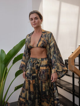 Woman modeling patterned resort wear with bikini top, flowing robe and wide leg pants in modern tropical interior