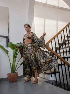 Woman modeling flowing patterned resort wear with bikini top and wide leg pants in bright modern interior
