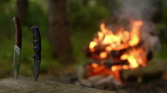 Outdoor bushcraft knives on log near warm burning campfire with natural crackling audio soundscape. Cinematic video background for wilderness survival gear, tactical knife review, camping.