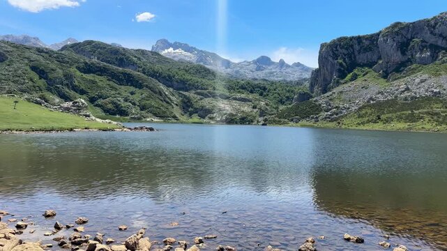 Breathtaking summer landscape of a serene mountain lake surrounded by lush green valleys, rocky peaks, and dramatic cloudy skies in Picos de Europa National Park, Spain. 