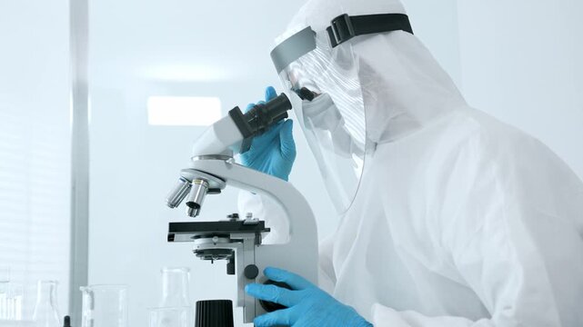 Scientist in a white cleanroom suit, face shield, and blue gloves conducting scientific research using a microscope in a laboratory, working with samples and developing medicine