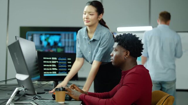 Young diverse colleagues collaborate in a modern IT control room. Asian female manager points at computer screen, discussing financial data charts with an African American male data analyst specialist