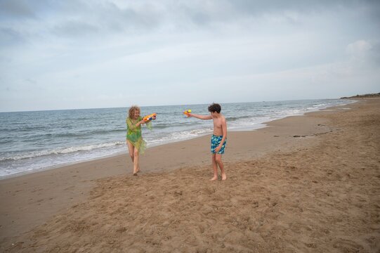 Mother and teenager son laughing while playfully squirting water guns at each other during a fun day together on the beach