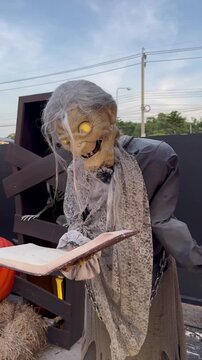 A spooky, elderly witch animatronic holding a mysterious book with glowing eyes, stands outside a haunted house during Halloween, turning itself from side to side, vertical video.