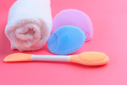 Different tools for skin care and oral care placed on a pink background with a towel and cleaning brushes