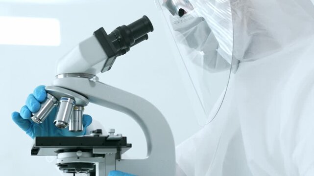Scientist in a white cleanroom suit, face shield, and blue gloves conducting scientific research using a microscope in a laboratory, working with samples and developing medicine