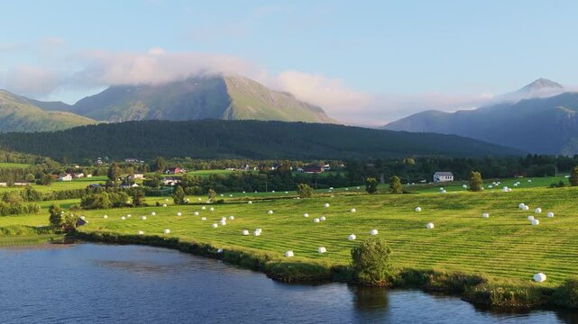 Aerial drone view of a fjord and hay bales in a field at sunset in Norway. Sunset glow on sky and clouds over mountain peaks during summer.