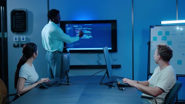 Young Diverse software engineering team collaborating in a modern tech office at night. Programmers coding and analyzing data on computers while a manager presents script on a large digital screen.