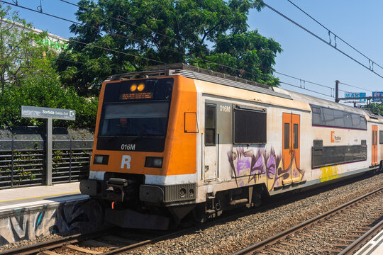 Barcelona, ​​Spain, June 29, 2026. A double-decker passenger train painted orange, white, and gray with graffiti on its side sits on train tracks next to a station exit sign.