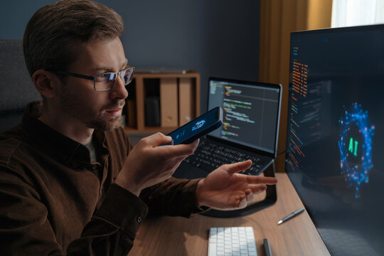 Close up of a man wearing glasses talking to an AI chatbot on a smartphone while working at a computer with artificial intelligence agent. Man using artificial intelligence assistant for work. 