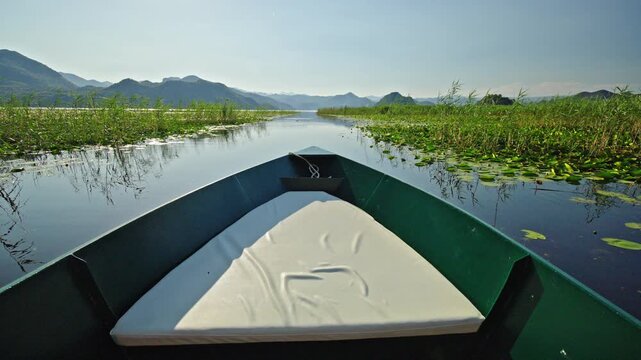 Scenic boat ride through the wetlands of Skadar Lake National Park in Montenegro. View from the bow of a traditional boat gliding between water lilies and reeds with mountains on the horizon.