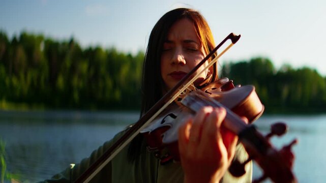Woman playing violin outdoors by a tranquil lakeside, eyes closed in concentration, expressing emotion through music against a peaceful forest and water backdrop