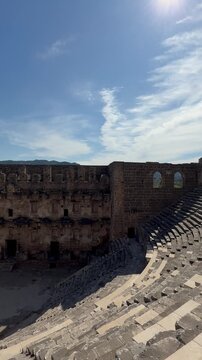 Camera pans left across ancient stone amphitheater interior, moving from close foreground masonry and worn walkway to reveal full semicircular stepped seating, carved risers, and central arena