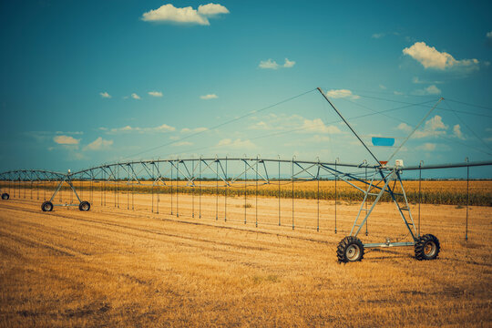 A wide agricultural landscape photo of a large center pivot irrigation system in a harvested golden field, styled with warm vintage film color grading and clear blue summer sky