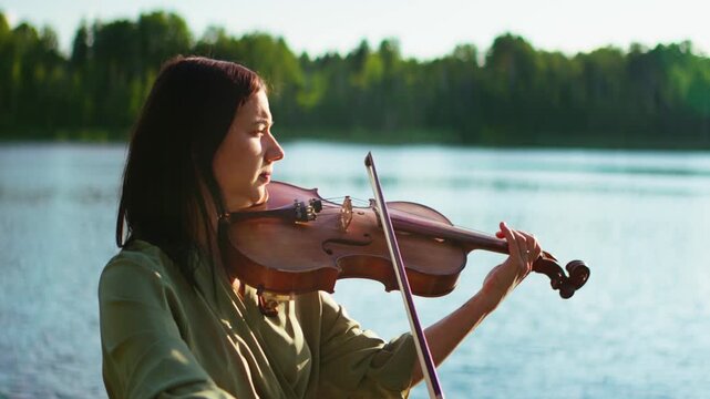 Woman playing violin outdoors by a tranquil lakeside at sunset, focusing on expressive classical music performance and peaceful connection with nature and water