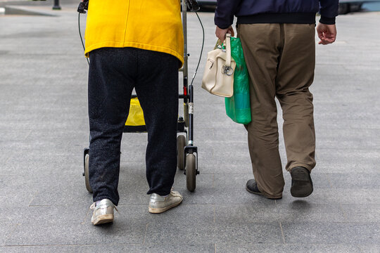 Two People Walking with a Mobility Walker and Shopping Bags. Rear view of two people walking on a paved plaza, one using a mobility walker and the other carrying shopping bags.