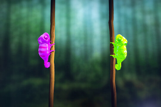 Bright Chameleon Figures on Vertical Branches. Two neon chameleon figures in pink and green cling to separate vertical branches against a softly blurred forest background.