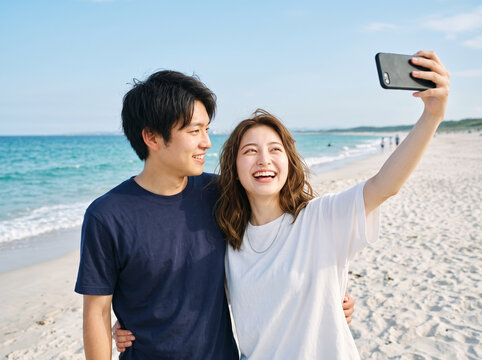 砂浜で紺Tシャツの男性に見守られながら満面の笑みで自撮り撮影する白Tシャツの女性