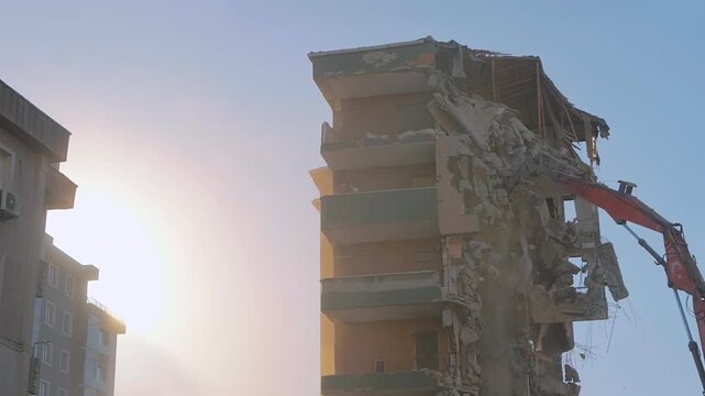 Excavator with crusher is tearing down the old typical Turkish multi-storey apartment, residential building in Istanbul, Turkey: demolition in slow motion. Setting sun is shining through the dust