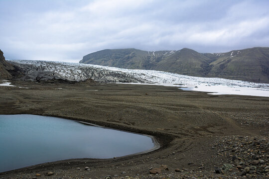 Vast Iceland landscape featuring a glacial tongue retreating from volcanic mountains, revealing a dark ground with a serene meltwater lake reflecting the cloudy sky