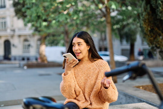 A young woman sits by a parked bicycle in an urban park, holding a smartphone and gazing thoughtfully into the distance. She wears a cozy sweater and jeans, suggesting a relaxed autumn outing. The can