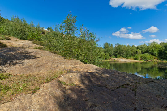 A scenic view of a tranquil lake bordered by green trees and a sandy bank, beneath a bright blue sky with clouds - Church Wood and Old Quarry, Bury, UK