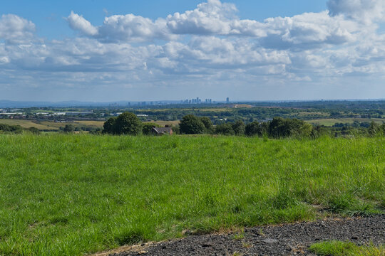 A bright landscape photo features a vibrant green field in the foreground. A distant city skyline is visible on the horizon beneath a cloudy sky - Church Wood and Old Quarry, Bury, UK