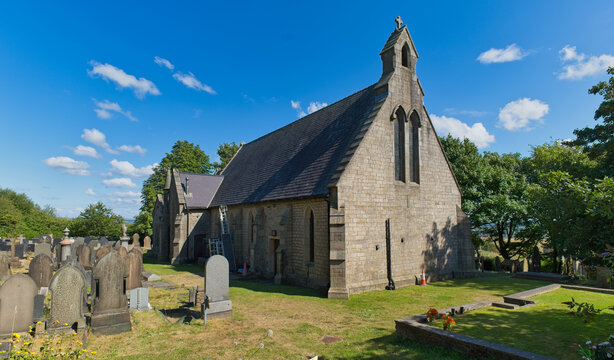 A weathered stone church with a steep roof stands in a sunny graveyard, surrounded by trees. The bright blue sky completes the serene scene - Church Wood and Old Quarry, Bury, UK