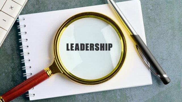 A magnifying glass shows the words leader and leadership on a notepad. The setting includes a pen and a light gray table. This captures ideas around leadership